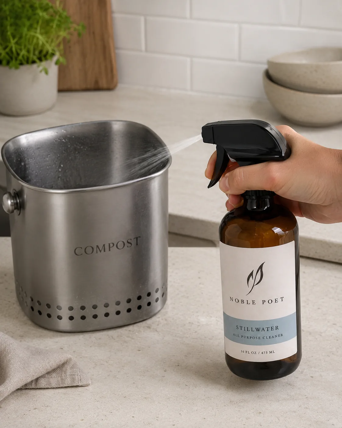 A hand spraying Noble Poet Stillwater into a stainless steel compost bin in a calm kitchen